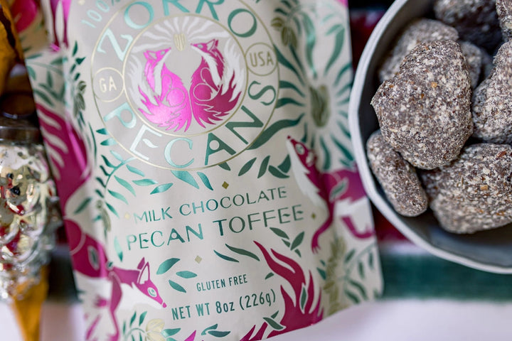 Zorro Milk Chocolate Pecan Toffee packaging beside a bowl of chocolate-coated pecan toffee pieces.